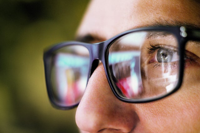 The picture shows a close-up of the eye area of a person wearing square, black glasses. The person is looking from the right side of the picture to the left. You can only see the person's left eye, which is grey-blue and surrounded by dark eyelashes. The focus of the picture is on the recognisable eye, the rest of the picture is blurry.