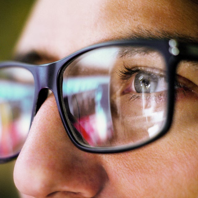 The picture shows a close-up of the eye area of a person wearing square, black glasses. The person is looking from the right side of the picture to the left. You can only see the person's left eye, which is grey-blue and surrounded by dark eyelashes. The focus of the picture is on the recognisable eye, the rest of the picture is blurry.