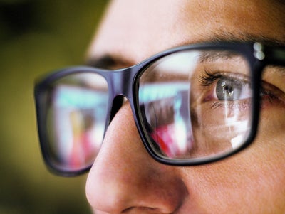 The picture shows a close-up of the eye area of a person wearing square, black glasses. The person is looking from the right side of the picture to the left. You can only see the person's left eye, which is grey-blue and surrounded by dark eyelashes. The focus of the picture is on the recognisable eye, the rest of the picture is blurry.