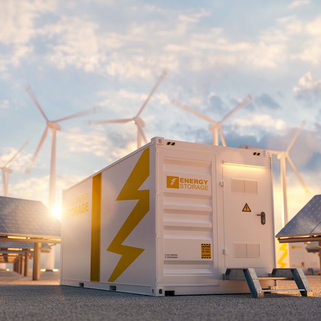 The picture shows a large white container in the centre, which is visible both from the side and from the front. On the top left-hand side of the container is orange lettering with the inscription ‘Energy Storage’. A large orange stripe runs from top to bottom in the centre of the side surface. To the right of this stripe is a large pictogram of a lightning bolt that extends from the top right to the bottom centre. The front of the container also features the words ‘Energy Storage’ on the left-hand side. To the right of this is a door with a black handle and a yellow warning triangle with a lightning bolt on it. As the door is not at ground level, a small metal staircase with two steps leads up to it. To the right and left of the container, several angled solar panels are lined up one behind the other. Several wind turbines can be seen in the background above the container, which are slightly blurred, indicating that they are in operation. The sky is slightly cloudy and the low sun indicates that it is later in the day, as it is already at the height of the container.