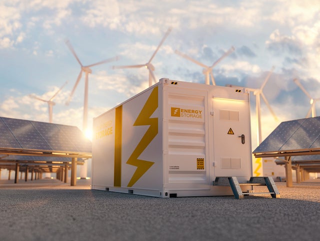 The picture shows a large white container in the centre, which is visible both from the side and from the front. On the top left-hand side of the container is orange lettering with the inscription ‘Energy Storage’. A large orange stripe runs from top to bottom in the centre of the side surface. To the right of this stripe is a large pictogram of a lightning bolt that extends from the top right to the bottom centre. The front of the container also features the words ‘Energy Storage’ on the left-hand side. To the right of this is a door with a black handle and a yellow warning triangle with a lightning bolt on it. As the door is not at ground level, a small metal staircase with two steps leads up to it. To the right and left of the container, several angled solar panels are lined up one behind the other. Several wind turbines can be seen in the background above the container, which are slightly blurred, indicating that they are in operation. The sky is slightly cloudy and the low sun indicates that it is later in the day, as it is already at the height of the container.