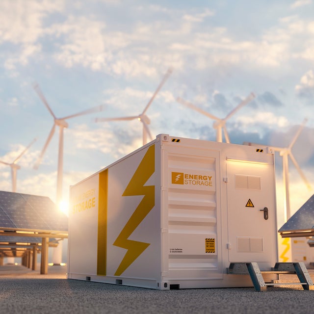 The picture shows a large white container in the centre, which is visible both from the side and from the front. On the top left-hand side of the container is orange lettering with the inscription ‘Energy Storage’. A large orange stripe runs from top to bottom in the centre of the side surface. To the right of this stripe is a large pictogram of a lightning bolt that extends from the top right to the bottom centre. The front of the container also features the words ‘Energy Storage’ on the left-hand side. To the right of this is a door with a black handle and a yellow warning triangle with a lightning bolt on it. As the door is not at ground level, a small metal staircase with two steps leads up to it. To the right and left of the container, several angled solar panels are lined up one behind the other. Several wind turbines can be seen in the background above the container, which are slightly blurred, indicating that they are in operation. The sky is slightly cloudy and the low sun indicates that it is later in the day, as it is already at the height of the container.