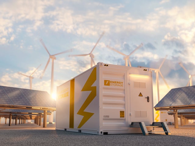 The picture shows a large white container in the centre, which is visible both from the side and from the front. On the top left-hand side of the container is orange lettering with the inscription ‘Energy Storage’. A large orange stripe runs from top to bottom in the centre of the side surface. To the right of this stripe is a large pictogram of a lightning bolt that extends from the top right to the bottom centre. The front of the container also features the words ‘Energy Storage’ on the left-hand side. To the right of this is a door with a black handle and a yellow warning triangle with a lightning bolt on it. As the door is not at ground level, a small metal staircase with two steps leads up to it. To the right and left of the container, several angled solar panels are lined up one behind the other. Several wind turbines can be seen in the background above the container, which are slightly blurred, indicating that they are in operation. The sky is slightly cloudy and the low sun indicates that it is later in the day, as it is already at the height of the container.