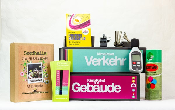 Auf dem Bild sieht man verschiedene Gegenstände vor einem weißen Hintergrund, die zu den Klimapaketen Verkehr und Gebäude gehören. Darunter zwei Boxen in den Farben Magenta und Türkis, sowie ein Messgerät, Pflanzensamen, eine Pannenhilfen Set für das Fahrrad u.v.m.