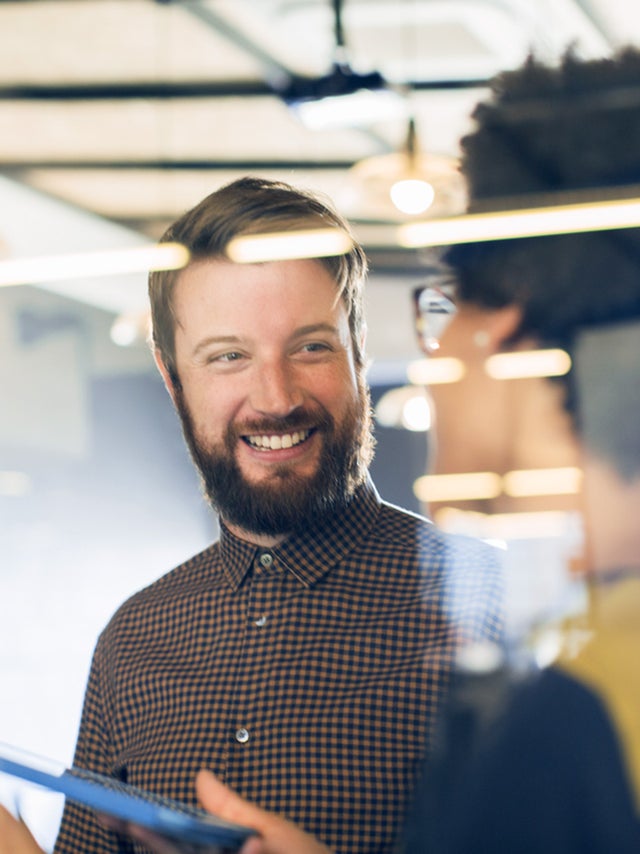 In the picture, a bearded man in a checkered shirt is smiling at a woman with curly hair and glasses who is facing him. The woman is holding a tablet in her right hand, and they appear to be in conversation. They are standing behind a glass partition. The setting is an office environment with ceiling lights visible in the background, and their reflections can be seen on the glass partition between them.