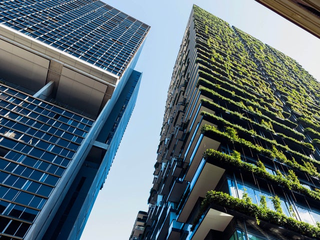 The picture shows a few skyscrapers that are seen from below. Two of them are clearly shown, the others are build behind them or can’t be seen from this point of view. On the skyscraper on the left side are just plain windows, but the skyscraper on the right side has greenery on every floor and is beeing illuminated by the sun.