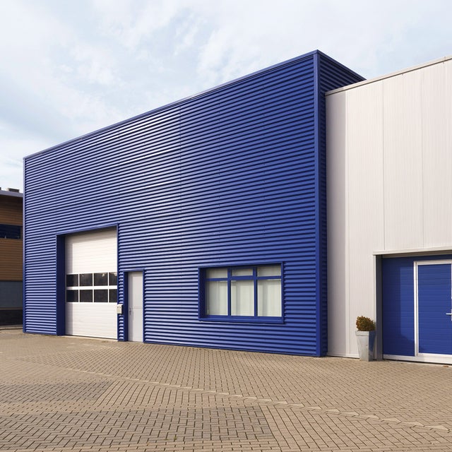 The picture shows a central warehouse with a horizontally fluted blue steel façade and a large white garage entrance for lorries on the left-hand side of the building. This entrance is decorated with eight windows. Next to the garage door is a simple white entrance door. On the right-hand side of the building, a window is divided into three sections and covered with white shutters on the inside. Attached to the warehouse on the right is a slightly lower, white fluted building, which also has a steel façade. The extension has a garage door at car height, which also contains a built-in entrance door. It is the same blue colour as the warehouse façade. To the left and right of this garage door are two taller pots with spherical plants. A drainage pipe runs along the white extension on the right-hand edge of the picture. In the left quarter of the picture, next to the blue warehouse, a modern house can be seen. It has an asymmetrical wood and stone façade decorated with windows of different sizes. In the lower third of the picture there is a kind of driveway and car park for the warehouse. The sky is bluish and slightly cloudy.