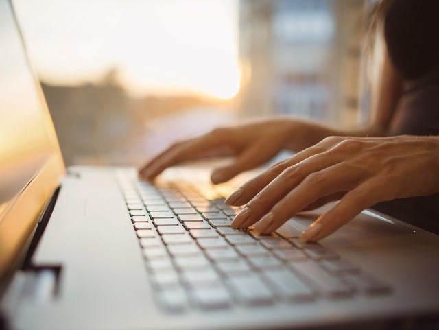 The picture shows a close-up of the keyboard of a laptop, which is illuminated by warm rays of sunlight from the background. The screen of the laptop is on the left edge of the picture and a woman in a dark blue shirt can be seen sitting in a blur on the right edge. Her hands are resting lightly on the keys of the laptop. The background of the picture is very blurred, but you can easily recognise the walls of a room and a window through which the sun's rays shine.