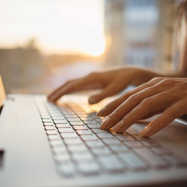 The picture shows a close-up of the keyboard of a laptop, which is illuminated by warm rays of sunlight from the background. The screen of the laptop is on the left edge of the picture and a woman in a dark blue shirt can be seen sitting in a blur on the right edge. Her hands are resting lightly on the keys of the laptop. The background of the picture is very blurred, but you can easily recognise the walls of a room and a window through which the sun's rays shine.