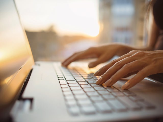 The picture shows a close-up of the keyboard of a laptop, which is illuminated by warm rays of sunlight from the background. The screen of the laptop is on the left edge of the picture and a woman in a dark blue shirt can be seen sitting in a blur on the right edge. Her hands are resting lightly on the keys of the laptop. The background of the picture is very blurred, but you can easily recognise the walls of a room and a window through which the sun's rays shine.