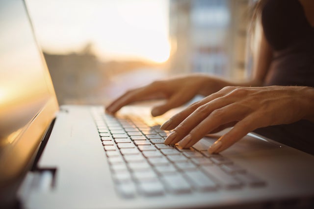 The picture shows a close-up of the keyboard of a laptop, which is illuminated by warm rays of sunlight from the background. The screen of the laptop is on the left edge of the picture and a woman in a dark blue shirt can be seen sitting in a blur on the right edge. Her hands are resting lightly on the keys of the laptop. The background of the picture is very blurred, but you can easily recognise the walls of a room and a window through which the sun's rays shine.