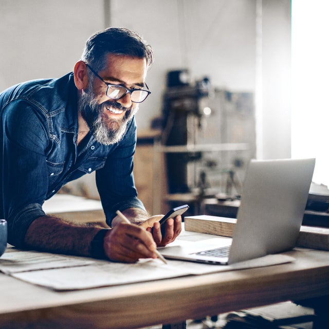 The picture shows a middle-aged man working in a workshop or studio. He has dark hair, wears glasses and his face is framed by a full, well-groomed grey beard. He wears a dark blue denim shirt and smiles broadly.The man is leaning over a large work table while holding a smartphone in his left hand looking at ista’s operating cost billing  and making notes on the paper with his right hand. There is an open laptop on the table in front of him. Next to the laptop is a blue coffee cup.The background of the workshop is blurred, but you can recognise shelves and tools that indicate a manual or creative activity. The light streaming in from the right through a window illuminates the scene and creates a warm, inviting atmosphere.The man appears focussed and content, which is conveyed by his smile and relaxed posture.