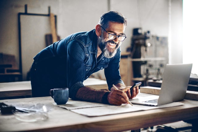 The picture shows a middle-aged man working in a workshop or studio. He has dark hair, wears glasses and his face is framed by a full, well-groomed grey beard. He wears a dark blue denim shirt and smiles broadly.The man is leaning over a large work table while holding a smartphone in his left hand looking at ista’s operating cost billing  and making notes on the paper with his right hand. There is an open laptop on the table in front of him. Next to the laptop is a blue coffee cup.The background of the workshop is blurred, but you can recognise shelves and tools that indicate a manual or creative activity. The light streaming in from the right through a window illuminates the scene and creates a warm, inviting atmosphere.The man appears focussed and content, which is conveyed by his smile and relaxed posture.