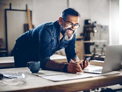 L'image montre un homme d'âge moyen travaillant dans un atelier ou un studio. Il a les cheveux noirs, porte des lunettes et son visage est encadré par une barbe grise fournie et bien entretenue. L'homme est penché sur une grande table de travail, tient un smartphone dans sa main gauche, regarde la facturation des coûts d'exploitation d'Ista et prend des notes sur le papier avec sa main droite. Un ordinateur portable ouvert se trouve sur la table devant lui. L'arrière-plan de l'atelier est flou, mais on peut reconnaître des étagères et des outils qui indiquent une activité manuelle ou créative. Le fond de l'atelier est flou, mais on peut reconnaître des étagères et des outils qui indiquent une activité manuelle ou créative. La lumière qui pénètre par la droite à travers une fenêtre éclaire la scène et crée une atmosphère chaleureuse et accueillante. L'homme semble concentré et satisfait, comme en témoignent son sourire et sa posture détendue.