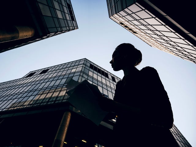 The picture shows a silhouette of a woman holding documents against the backdrop of modern skyscrapers. The perspective of the picture is from the bottom up, making the buildings look monumental and impressive. The woman is in the foreground of the picture, turned slightly to the left. Her profile is clearly visible and she appears to be looking intently at her documents. She is wearing a jacket and has tied her hair up in a bun. Several glass buildings can be seen in the background, taking up most of the picture. The building on the left of the picture is only partially visible, with a large round pillar extending upwards. The centre building is the most dominant and extends almost to the top of the picture. It has a glass and steel façade and reflects the sky. Another building can be seen on the right of the picture, which also extends upwards and forms a right angle to the centre building. The glass façades of the buildings give the picture a modern and urban feel. The sky is clear and slightly bluish, indicating a sunny or at least clear day. 