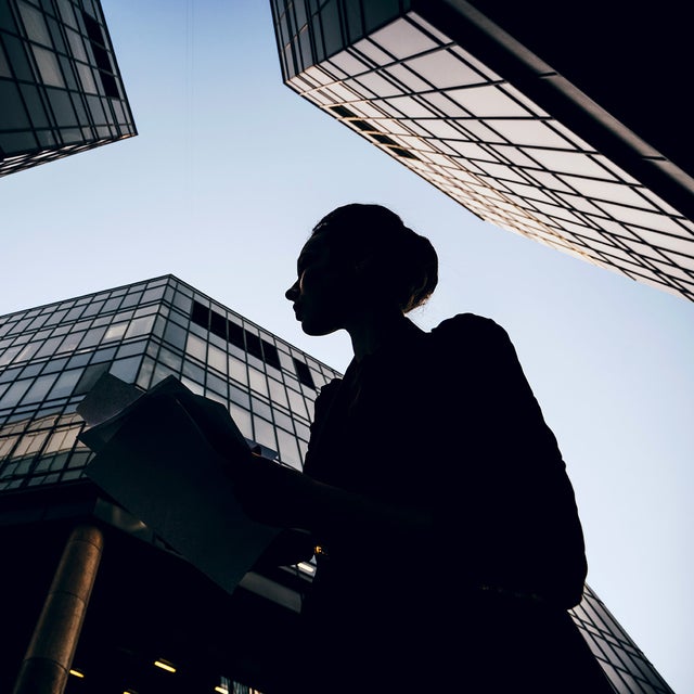 The picture shows a silhouette of a woman holding documents against the backdrop of modern skyscrapers. The perspective of the picture is from the bottom up, making the buildings look monumental and impressive. The woman is in the foreground of the picture, turned slightly to the left. Her profile is clearly visible and she appears to be looking intently at her documents. She is wearing a jacket and has tied her hair up in a bun. Several glass buildings can be seen in the background, taking up most of the picture. The building on the left of the picture is only partially visible, with a large round pillar extending upwards. The centre building is the most dominant and extends almost to the top of the picture. It has a glass and steel façade and reflects the sky. Another building can be seen on the right of the picture, which also extends upwards and forms a right angle to the centre building. The glass façades of the buildings give the picture a modern and urban feel. The sky is clear and slightly bluish, indicating a sunny or at least clear day. 