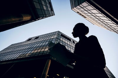 The picture shows a silhouette of a woman holding documents against the backdrop of modern skyscrapers. The perspective of the picture is from the bottom up, making the buildings look monumental and impressive. The woman is in the foreground of the picture, turned slightly to the left. Her profile is clearly visible and she appears to be looking intently at her documents. She is wearing a jacket and has tied her hair up in a bun. Several glass buildings can be seen in the background, taking up most of the picture. The building on the left of the picture is only partially visible, with a large round pillar extending upwards. The centre building is the most dominant and extends almost to the top of the picture. It has a glass and steel façade and reflects the sky. Another building can be seen on the right of the picture, which also extends upwards and forms a right angle to the centre building. The glass façades of the buildings give the picture a modern and urban feel. The sky is clear and slightly bluish, indicating a sunny or at least clear day. 