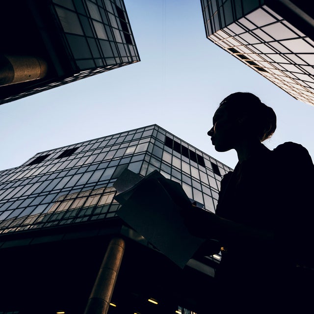 The picture shows a silhouette of a woman holding documents against the backdrop of modern skyscrapers. The perspective of the picture is from the bottom up, making the buildings look monumental and impressive. The woman is in the foreground of the picture, turned slightly to the left. Her profile is clearly visible and she appears to be looking intently at her documents. She is wearing a jacket and has tied her hair up in a bun. Several glass buildings can be seen in the background, taking up most of the picture. The building on the left of the picture is only partially visible, with a large round pillar extending upwards. The centre building is the most dominant and extends almost to the top of the picture. It has a glass and steel façade and reflects the sky. Another building can be seen on the right of the picture, which also extends upwards and forms a right angle to the centre building. The glass façades of the buildings give the picture a modern and urban feel. The sky is clear and slightly bluish, indicating a sunny or at least clear day. 