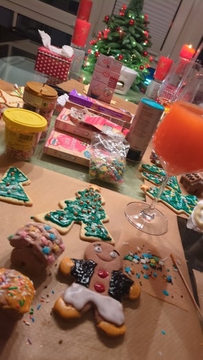 Ein Tisch mit Weihnachtskeksen in verschiedenen Formen, wie Tannenbäume und Lebkuchenmännchen, dekoriert mit buntem Zuckerguss und Streuseln. Im Hintergrund stehen Weihnachtsdekorationen, darunter ein kleiner beleuchteter Weihnachtsbaum und rote Kerzen.