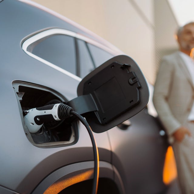 The picture shows an electric car that is being charged. In the foreground is the car's charging socket, into which a charging cable is plugged. The cable is connected to an ista EV charging station, which is outside the picture. In the background, slightly out of focus, is a man in a light-coloured suit talking on the phone. He has a relaxed posture and is leaning slightly against the car. The scene probably takes place in an urban environment or in a company car park, which is indicated by the modern buildings in the background. The image conveys an atmosphere of modern technology and environmentally friendly mobility.