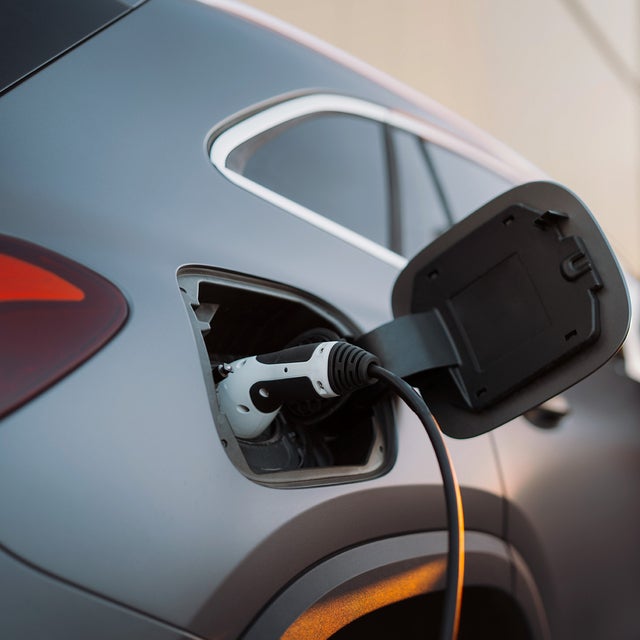 The picture shows an electric car that is being charged. In the foreground is the car's charging socket, into which a charging cable is plugged. The cable is connected to an ista EV charging station, which is outside the picture. In the background, slightly out of focus, is a man in a light-coloured suit talking on the phone. He has a relaxed posture and is leaning slightly against the car. The scene probably takes place in an urban environment or in a company car park, which is indicated by the modern buildings in the background. The image conveys an atmosphere of modern technology and environmentally friendly mobility.