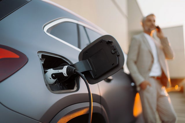 The picture shows an electric car that is being charged. In the foreground is the car's charging socket, into which a charging cable is plugged. The cable is connected to an ista EV charging station, which is outside the picture. In the background, slightly out of focus, is a man in a light-coloured suit talking on the phone. He has a relaxed posture and is leaning slightly against the car. The scene probably takes place in an urban environment or in a company car park, which is indicated by the modern buildings in the background. The image conveys an atmosphere of modern technology and environmentally friendly mobility.