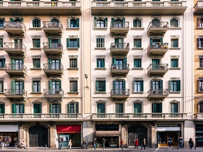 Frontalansicht von 3 Reihenhäusern im urbanen Umfeld. Jede Front ist in einem hellem Farbton gestrichen und es sind zahlreiche Fenster und je zwei Balkone pro Stockwerk zu erkennen.  Unten in den Gebäuden sind Geschäfte die ihre Markisen ausgefahren haben. Mehrere Menschen sind auf der Straße unterwegs. 