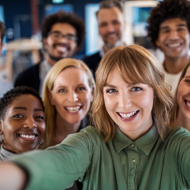 The image shows a group of diverse people smiling and taking a group selfie. They are standing close together in an indoor setting that appears to be an office or a collaborative workspace. The people in the photo represent a variety of ages, genders, and ethnic backgrounds, reflecting a diverse and inclusive environment. The atmosphere is cheerful and positive, suggesting a sense of camaraderie and teamwork among the group members.