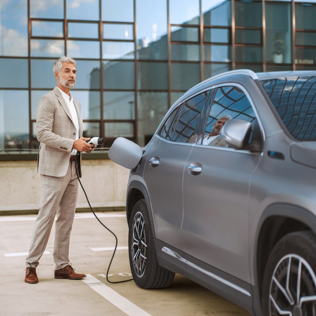 The picture shows an electric car that is being charged. In the foreground is the car's charging socket, into which a charging cable is plugged. The cable is connected to an ista EV charging station, which is outside the picture. In the background, slightly out of focus, is a man in a light-coloured suit talking on the phone. He has a relaxed posture and is leaning slightly against the car. The scene probably takes place in an urban environment or in a company car park, which is indicated by the modern buildings in the background. The image conveys an atmosphere of modern technology and environmentally friendly mobility.