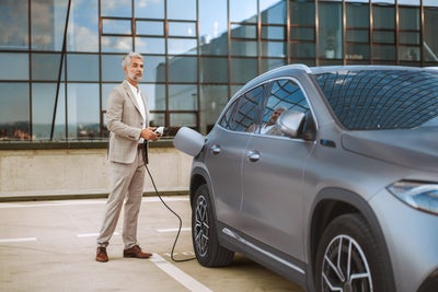 The picture shows an electric car that is being charged. In the foreground is the car's charging socket, into which a charging cable is plugged. The cable is connected to an ista EV charging station, which is outside the picture. In the background, slightly out of focus, is a man in a light-coloured suit talking on the phone. He has a relaxed posture and is leaning slightly against the car. The scene probably takes place in an urban environment or in a company car park, which is indicated by the modern buildings in the background. The image conveys an atmosphere of modern technology and environmentally friendly mobility.