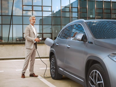 The picture shows an electric car that is being charged. In the foreground is the car's charging socket, into which a charging cable is plugged. The cable is connected to an ista EV charging station, which is outside the picture. In the background, slightly out of focus, is a man in a light-coloured suit talking on the phone. He has a relaxed posture and is leaning slightly against the car. The scene probably takes place in an urban environment or in a company car park, which is indicated by the modern buildings in the background. The image conveys an atmosphere of modern technology and environmentally friendly mobility.