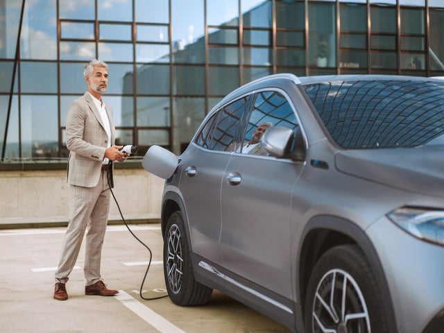 The picture shows an electric car that is being charged. In the foreground is the car's charging socket, into which a charging cable is plugged. The cable is connected to an ista EV charging station, which is outside the picture. In the background, slightly out of focus, is a man in a light-coloured suit talking on the phone. He has a relaxed posture and is leaning slightly against the car. The scene probably takes place in an urban environment or in a company car park, which is indicated by the modern buildings in the background. The image conveys an atmosphere of modern technology and environmentally friendly mobility.