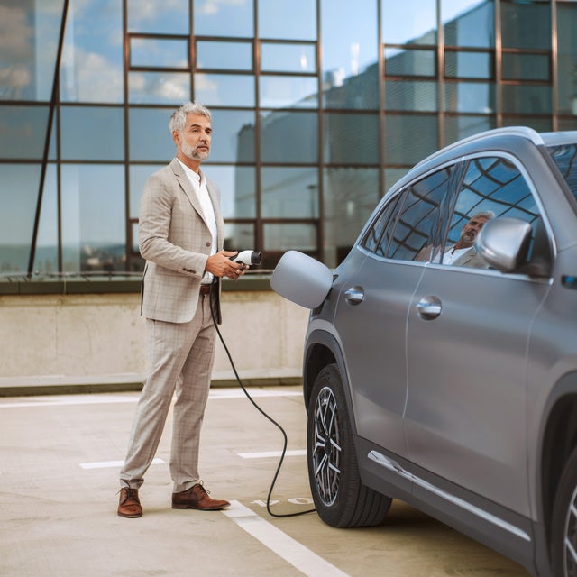 The picture shows an electric car that is being charged. In the foreground is the car's charging socket, into which a charging cable is plugged. The cable is connected to an ista EV charging station, which is outside the picture. In the background, slightly out of focus, is a man in a light-coloured suit talking on the phone. He has a relaxed posture and is leaning slightly against the car. The scene probably takes place in an urban environment or in a company car park, which is indicated by the modern buildings in the background. The image conveys an atmosphere of modern technology and environmentally friendly mobility.