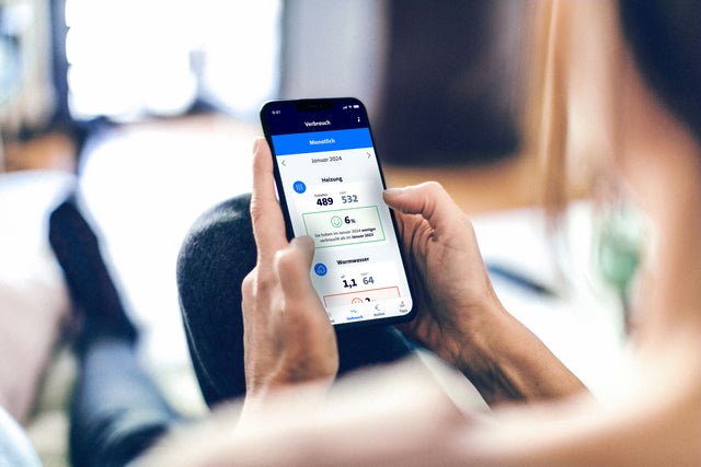 Man blickt aus Nutzerperspektive auf den Bildschirm eines Smartphones, welche mit beiden Händen gehalten wird. Auf dem Bildschirm erkennt man die monatliche Verbrauchsinformation aus EcoTrend. Abgebildet ist der Januar 2024, die Nutzerin konnte ihre Heizverbrauch im Vergleich zum Vorjahr um 6% verbessern.  Es scheint jedoch eine Verschlechterung bei Warmwasser gegeben zu haben. 