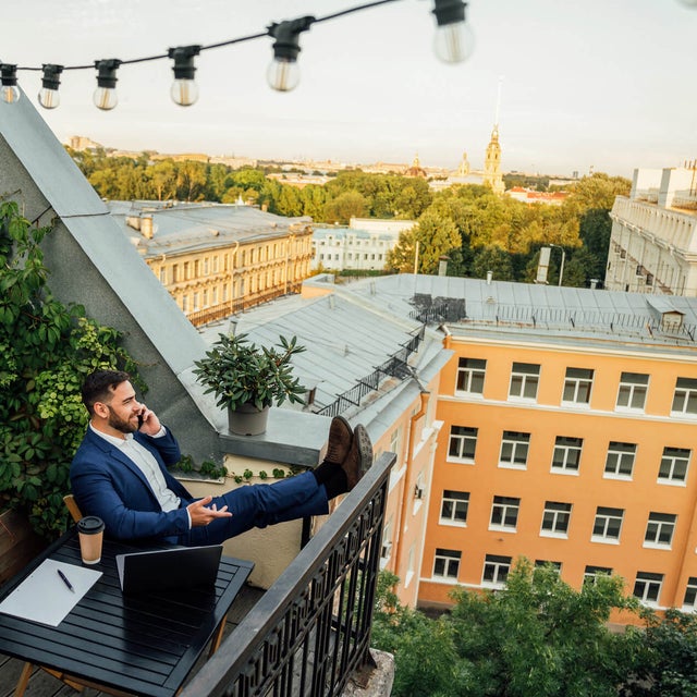 The picture shows a man sitting on a balcony in an urban environment. He is wearing a dark blue suit and a white shirt and is sitting relaxed at a small black table on the lower half in the centre of the picture. On the table in front of him are a laptop, a notepad with a pen and a coffee mug. The man has rested his legs on the balcony railing and is holding a telephone in his left hand while talking on the phone. The balcony is richly decorated with plants. On the left of the picture, climbing plants stretch up the wall, taking up almost the entire left side of the picture. Below the climbing plants are several pots of flowering plants and greenery arranged along the floor of the balcony. Above the man, stretched across the balcony, is a string of lights with several light bulbs that extends from the top left-hand corner to the right-hand side of the picture. In the background you can see a row of multi-storey buildings in various shades of yellow stretching across the top and centre of the picture. Behind the buildings is a park-like landscape with many trees that further break up the background. The sun seems to be either rising or setting, giving the picture a warm, golden glow. The view from the balcony extends over the roofs of the city.