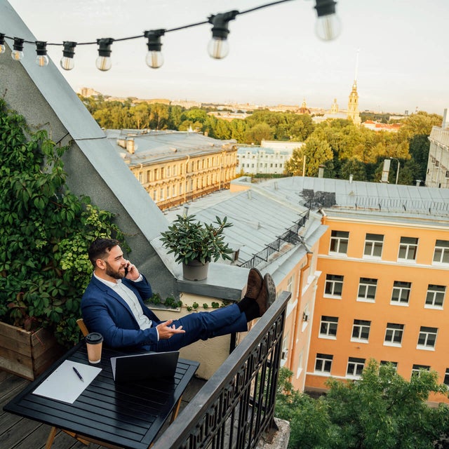 The picture shows a man sitting on a balcony in an urban environment. He is wearing a dark blue suit and a white shirt and is sitting relaxed at a small black table on the lower half in the centre of the picture. On the table in front of him are a laptop, a notepad with a pen and a coffee mug. The man has rested his legs on the balcony railing and is holding a telephone in his left hand while talking on the phone. The balcony is richly decorated with plants. On the left of the picture, climbing plants stretch up the wall, taking up almost the entire left side of the picture. Below the climbing plants are several pots of flowering plants and greenery arranged along the floor of the balcony. Above the man, stretched across the balcony, is a string of lights with several light bulbs that extends from the top left-hand corner to the right-hand side of the picture. In the background you can see a row of multi-storey buildings in various shades of yellow stretching across the top and centre of the picture. Behind the buildings is a park-like landscape with many trees that further break up the background. The sun seems to be either rising or setting, giving the picture a warm, golden glow. The view from the balcony extends over the roofs of the city.