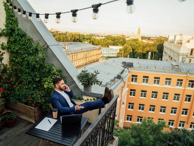 The picture shows a man sitting on a balcony in an urban environment. He is wearing a dark blue suit and a white shirt and is sitting relaxed at a small black table on the lower half in the centre of the picture. On the table in front of him are a laptop, a notepad with a pen and a coffee mug. The man has rested his legs on the balcony railing and is holding a telephone in his left hand while talking on the phone. The balcony is richly decorated with plants. On the left of the picture, climbing plants stretch up the wall, taking up almost the entire left side of the picture. Below the climbing plants are several pots of flowering plants and greenery arranged along the floor of the balcony. Above the man, stretched across the balcony, is a string of lights with several light bulbs that extends from the top left-hand corner to the right-hand side of the picture. In the background you can see a row of multi-storey buildings in various shades of yellow stretching across the top and centre of the picture. Behind the buildings is a park-like landscape with many trees that further break up the background. The sun seems to be either rising or setting, giving the picture a warm, golden glow. The view from the balcony extends over the roofs of the city.