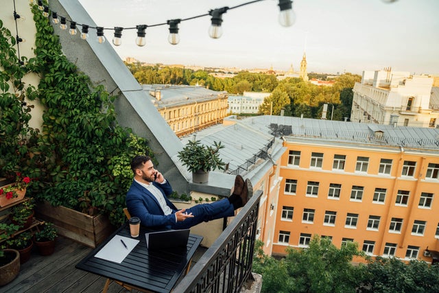 The picture shows a man sitting on a balcony in an urban environment. He is wearing a dark blue suit and a white shirt and is sitting relaxed at a small black table on the lower half in the centre of the picture. On the table in front of him are a laptop, a notepad with a pen and a coffee mug. The man has rested his legs on the balcony railing and is holding a telephone in his left hand while talking on the phone. The balcony is richly decorated with plants. On the left of the picture, climbing plants stretch up the wall, taking up almost the entire left side of the picture. Below the climbing plants are several pots of flowering plants and greenery arranged along the floor of the balcony. Above the man, stretched across the balcony, is a string of lights with several light bulbs that extends from the top left-hand corner to the right-hand side of the picture. In the background you can see a row of multi-storey buildings in various shades of yellow stretching across the top and centre of the picture. Behind the buildings is a park-like landscape with many trees that further break up the background. The sun seems to be either rising or setting, giving the picture a warm, golden glow. The view from the balcony extends over the roofs of the city.