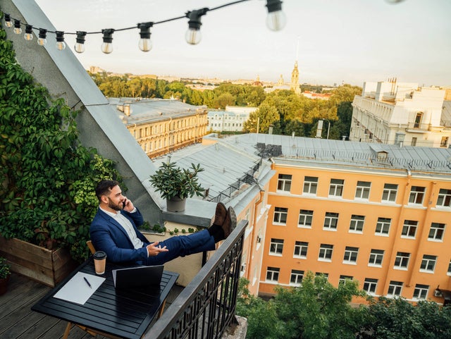 The picture shows a man sitting on a balcony in an urban environment. He is wearing a dark blue suit and a white shirt and is sitting relaxed at a small black table on the lower half in the centre of the picture. On the table in front of him are a laptop, a notepad with a pen and a coffee mug. The man has rested his legs on the balcony railing and is holding a telephone in his left hand while talking on the phone. The balcony is richly decorated with plants. On the left of the picture, climbing plants stretch up the wall, taking up almost the entire left side of the picture. Below the climbing plants are several pots of flowering plants and greenery arranged along the floor of the balcony. Above the man, stretched across the balcony, is a string of lights with several light bulbs that extends from the top left-hand corner to the right-hand side of the picture. In the background you can see a row of multi-storey buildings in various shades of yellow stretching across the top and centre of the picture. Behind the buildings is a park-like landscape with many trees that further break up the background. The sun seems to be either rising or setting, giving the picture a warm, golden glow. The view from the balcony extends over the roofs of the city.