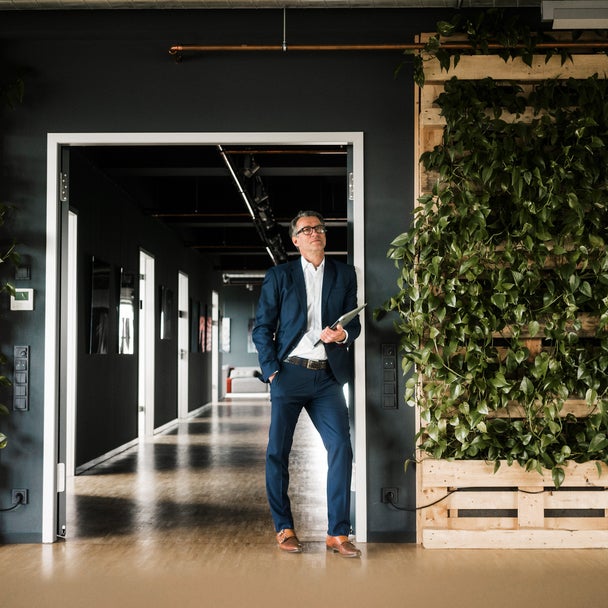 The picture shows a man with short grey hair and glasses in focus. He is wearing a blue suit with a white shirt and brown shoes. While his right hand is in his trouser pocket, he is holding a closed laptop in his other hand. He is leaning against a door frame that leads to a corridor in the background. Several doors can be seen on the left-hand side of the corridor, indicating various offices. To the right and left of the man on the walls are several vertically stacked pallets with green plants running down the pallets, giving the building a modern atmosphere.