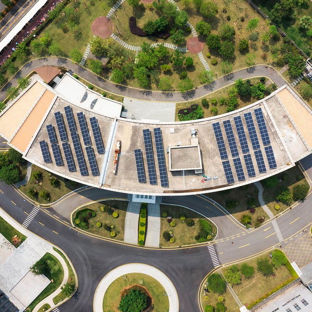 The image shows an aerial view of a modern building complex surrounded by streets, car parks and green spaces. In the centre of the image is a large, curved building with a roof surface that is completely covered with solar panels. The solar panels are arranged in several parallel rows and utilise the entire roof surface. Below the curved building is a wide road that winds around a circular green area with a tree in the centre. This road separates the main building from another large, rectangular building, which is located in the lower left-hand section of the picture. This building has a flat roof and is surrounded by grey paved areas and other green spaces. At the top of the picture is an extensive park with various footpaths running through it. There are numerous trees and shrubs in the park, creating a natural and green environment. At the top right of the picture is a long, straight road that separates the park area from the rest of the grounds. Another road is visible on the left-hand side of the picture, which also has a cycle path and footpath to the left and right of the road. Yellow markings show an entrance to the building complex which is blocked by a barrier.