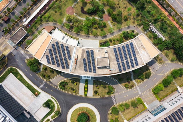 The image shows an aerial view of a modern building complex surrounded by streets, car parks and green spaces. In the centre of the image is a large, curved building with a roof surface that is completely covered with solar panels. The solar panels are arranged in several parallel rows and utilise the entire roof surface. Below the curved building is a wide road that winds around a circular green area with a tree in the centre. This road separates the main building from another large, rectangular building, which is located in the lower left-hand section of the picture. This building has a flat roof and is surrounded by grey paved areas and other green spaces. At the top of the picture is an extensive park with various footpaths running through it. There are numerous trees and shrubs in the park, creating a natural and green environment. At the top right of the picture is a long, straight road that separates the park area from the rest of the grounds. Another road is visible on the left-hand side of the picture, which also has a cycle path and footpath to the left and right of the road. Yellow markings show an entrance to the building complex which is blocked by a barrier.