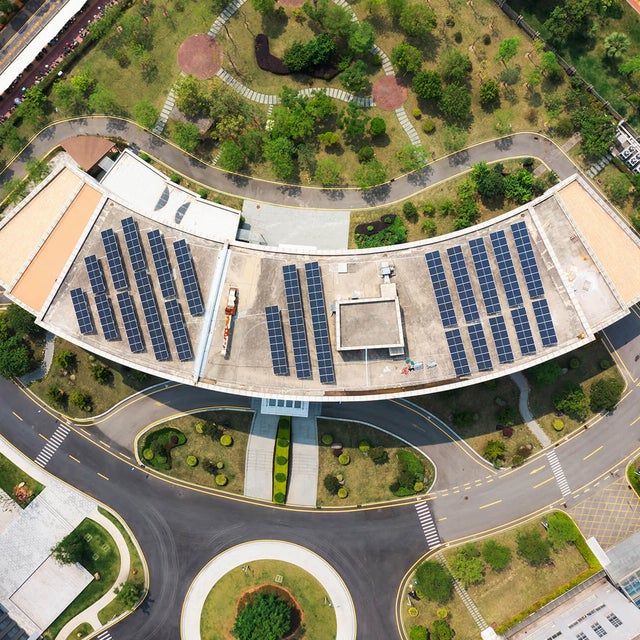 The image shows an aerial view of a modern building complex surrounded by streets, car parks and green spaces. In the centre of the image is a large, curved building with a roof surface that is completely covered with solar panels. The solar panels are arranged in several parallel rows and utilise the entire roof surface. Below the curved building is a wide road that winds around a circular green area with a tree in the centre. This road separates the main building from another large, rectangular building, which is located in the lower left-hand section of the picture. This building has a flat roof and is surrounded by grey paved areas and other green spaces. At the top of the picture is an extensive park with various footpaths running through it. There are numerous trees and shrubs in the park, creating a natural and green environment. At the top right of the picture is a long, straight road that separates the park area from the rest of the grounds. Another road is visible on the left-hand side of the picture, which also has a cycle path and footpath to the left and right of the road. Yellow markings show an entrance to the building complex which is blocked by a barrier.