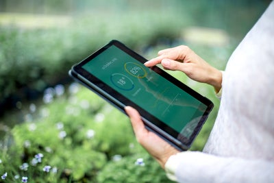 In the picture, a person is holding a tablet with both hands. The tablet screen displays data with percentages and graphs, including an 82% humidity reading and an 18°C temperature. The person is using their right index finger to interact with the screen. The background shows a blurred, green, outdoor environment with small white flowers.