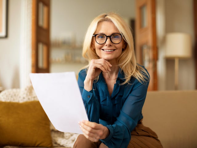 The picture shows a woman sitting on a couch and reading the ista heating bill. She has long blonde hair and is wearing black glasses. Her top is a blue shirt and she is wearing brown trousers. While she holds the heating bill in her left hand, her right hand rests on her chin. She looks directly into the camera. The beige couch on which she is sitting stretches across the entire lower third of the picture and has a white blanket draped over the back on the left with a yellow cushion leaning against it. In the background are various pieces of furniture that suggest a living room: A floor lamp and a shelf can be seen on the right, and in the centre an open two-part door leading into another room with bookshelves. The entire background is blurred.