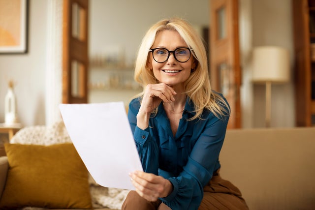 L'image montre une femme assise sur un canapé et lisant la facture de chauffage ista. Elle a de longs cheveux blonds et porte des lunettes noires. Son haut est une chemise bleue et elle porte un pantalon marron. Alors qu'elle tient la facture de chauffage dans sa main gauche, sa main droite est posée sur son menton. Elle regarde directement la caméra. Le canapé beige sur lequel elle est assise s'étend sur tout le tiers inférieur de l'image et comporte une couverture blanche drapée sur le dossier à gauche, ainsi qu'un coussin jaune adossé à celui-ci. À l'arrière-plan, divers meubles évoquent un salon : À droite, on aperçoit un lampadaire et une étagère, et au centre, une porte ouverte en deux parties donnant accès à une autre pièce avec des étagères. L'ensemble de l'arrière-plan est flou.