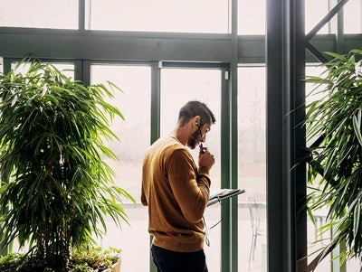 In the picture, a man wearing a mustard-colored sweater stands indoors near a large window. He is holding a pen and a notebook, appearing to be in deep thought or contemplation. He has earphones in his ears, suggesting he might be on a call or listening to something. On the left and right sides of the man, there are large green plants, adding a natural element to the indoor setting. The background shows an outdoor scene through the window, with faint outlines of trees and a patio area.