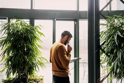 In the picture, a man wearing a mustard-colored sweater stands indoors near a large window. He is holding a pen and a notebook, appearing to be in deep thought or contemplation. He has earphones in his ears, suggesting he might be on a call or listening to something. On the left and right sides of the man, there are large green plants, adding a natural element to the indoor setting. The background shows an outdoor scene through the window, with faint outlines of trees and a patio area.