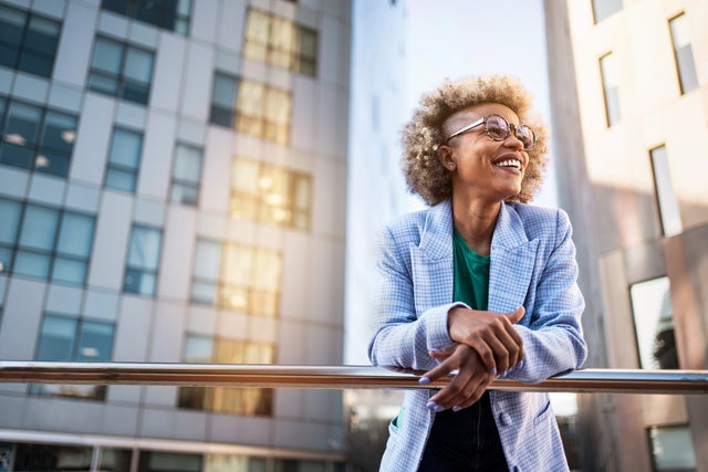 The picture shows a young woman standing on a raised platform or balcony. She is leaning casually over a shiny metal railing and smiling happily. The woman has blonde, curly hair with a fashionable undercut and wears large, round glasses. She is dressed in a light blue chequered blazer and a green T-shirt. In the background you can see modern office buildings with lots of windows. These buildings are grey and reflect the sunlight, giving the picture a dynamic and lively atmosphere. The light comes from the right-hand side and creates bright reflections on the windows and the railings. The woman is in the centre of the picture, leaning slightly to the right as she grasps the railing. Her facial expression is relaxed and content, as if she is enjoying the view and the moment. The setting suggests an urban location, probably in a city with modern architectural elements.