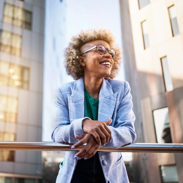 The picture shows a young woman standing on a raised platform or balcony. She is leaning casually over a shiny metal railing and smiling happily. The woman has blonde, curly hair with a fashionable undercut and wears large, round glasses. She is dressed in a light blue chequered blazer and a green T-shirt. In the background you can see modern office buildings with lots of windows. These buildings are grey and reflect the sunlight, giving the picture a dynamic and lively atmosphere. The light comes from the right-hand side and creates bright reflections on the windows and the railings. The woman is in the centre of the picture, leaning slightly to the right as she grasps the railing. Her facial expression is relaxed and content, as if she is enjoying the view and the moment. The setting suggests an urban location, probably in a city with modern architectural elements.